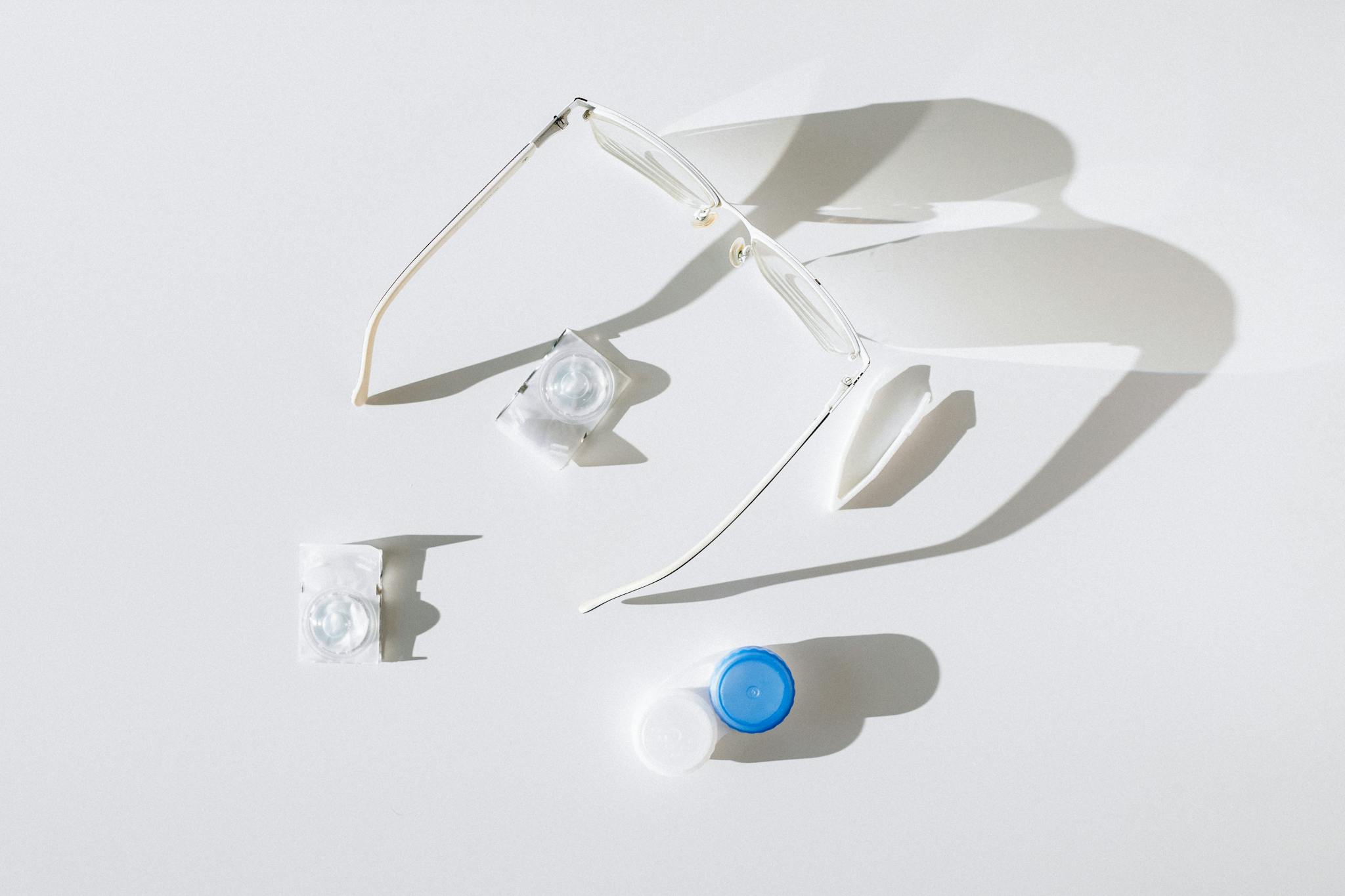 Flat lay of eyeglasses, contact lenses, and case on white background, emphasizing vision care.
